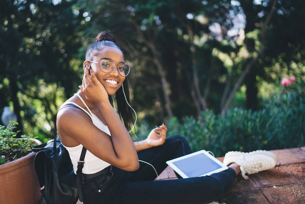 Portrait of cheerful female blogger in spectacles for vision protection smiling at camera while listening music playlist on leisure, happy African American woman in headphones using touch pad app - Zdjęcie, obraz