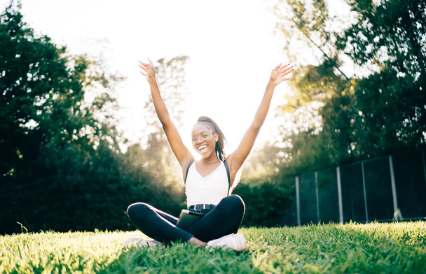 Portrait of cheerful hipster girl in trendy spectacles for provide eyes correction raising hands in air and rejoicing during positive pastime in park, joyful female smiling at camera resting on grass - Photo, image