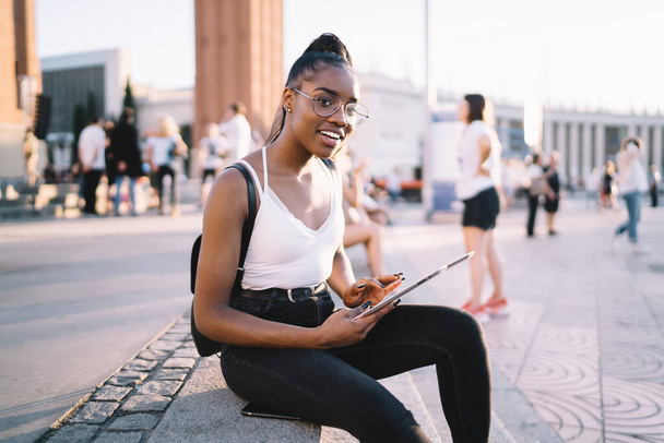 Portrait of happy hipster girl in stylish eyeglasses using touch pad device during travel vacations, happy dark skinned tourist with digital technology smiling at camera during time for rest in city - Valokuva, kuva