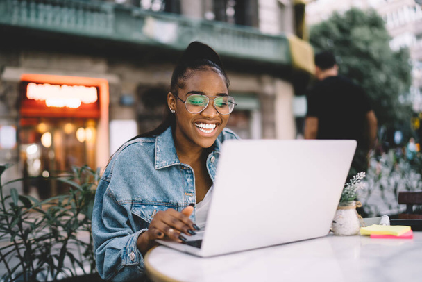 Joyful dark skinned female blogger typing text of publication using laptop computer working remotely, cheerful African American woman in spectacles share positive content and media files on netbook - Φωτογραφία, εικόνα