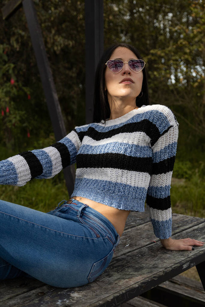 full body of young latin woman with straight black hair sitting relaxed on a park bench, wearing sunglasses and casual clothes, beauty and fashion - 写真・画像