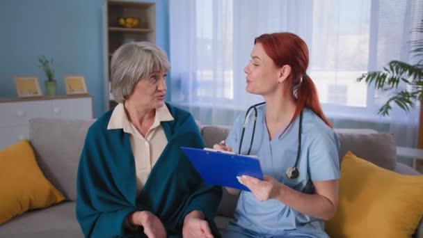 улыбающаяся медсестра обнимает плечи счастливой пациентки и смотрит в камеру, приятный доктор делает заметки на планшете и разговаривает с пожилой женщиной дома - Кадры, видео