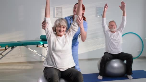 physiotherapist in uniform helping elderly people to do exercise with hands, old gray-haired couple on fitballs looking at camera with doctor - Záběry, video