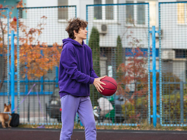 Cute teenager in violet hoodie playing basketball. Young boy with ball learning dribble and shooting on the city court. Hobby for kids, active lifestyle - Foto, imagen