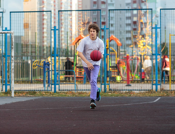 Cute teenager in a white t-shirt playing basketball outside. Young boy with ball learning dribble and shooting on the city court. Hobby for kids, active lifestyle - Фото, изображение