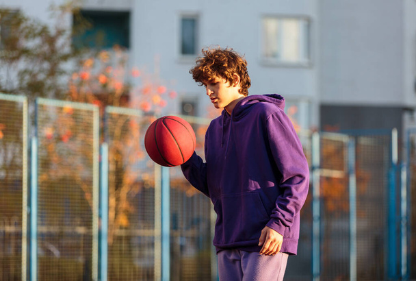 Χαριτωμένος έφηβος με μωβ κουκούλα που παίζει μπάσκετ. Νεαρό αγόρι με μπάλα μαθαίνει ντρίμπλα και σουτάρει στο γήπεδο της πόλης. Χόμπι για παιδιά, ενεργός τρόπος ζωής - Φωτογραφία, εικόνα
