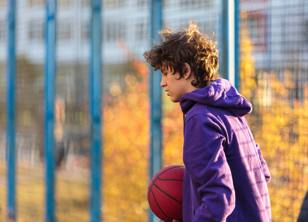 Adolescente carina con cappuccio viola che gioca a basket. Ragazzo con palla imparare dribblare e sparare sul campo della città. Hobby per bambini, stile di vita attivo - Foto, immagini