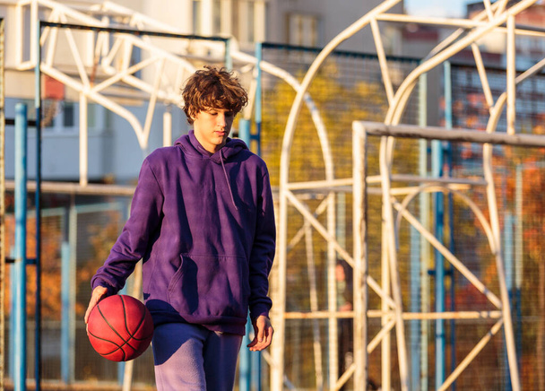 Cute teenager in violet hoodie playing basketball. Young boy with ball learning dribble and shooting on the city court. Hobby for kids, active lifestyle - Photo, Image