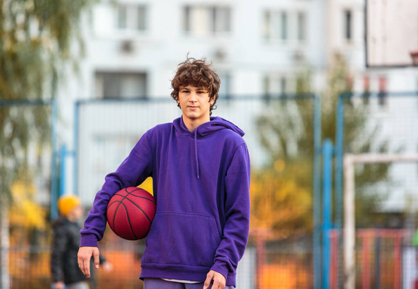 Χαριτωμένος έφηβος με μωβ κουκούλα που παίζει μπάσκετ. Νεαρό αγόρι με μπάλα μαθαίνει ντρίμπλα και σουτάρει στο γήπεδο της πόλης. Χόμπι για παιδιά, ενεργός τρόπος ζωής - Φωτογραφία, εικόνα