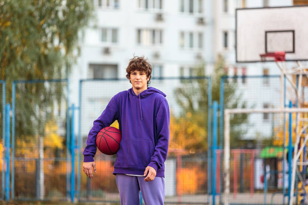 Netter Teenager in violettem Kapuzenpulli beim Basketballspielen. Junge mit Ball lernen Dribbeln und Schießen auf dem Stadtplatz. Hobby für Kinder, aktiver Lebensstil - Foto, Bild