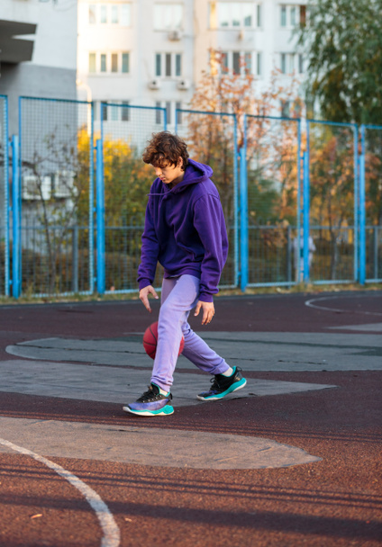 Cute teenager in violet hoodie playing basketball. Young boy with ball learning dribble and shooting on the city court. Hobby for kids, active lifestyle - Fotografie, Obrázek