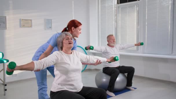 vreugdevol senior koppel zittend op oefenballen steek handen op met halters tijdens de training samen met verpleegster thuis - Video