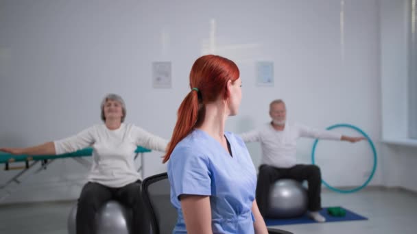 спорт и здоровый образ жизни, портрет счастливой женщины-врача в синей форме на фоне пожилой пары, занимающейся гимнастикой на мячах для упражнений - Кадры, видео