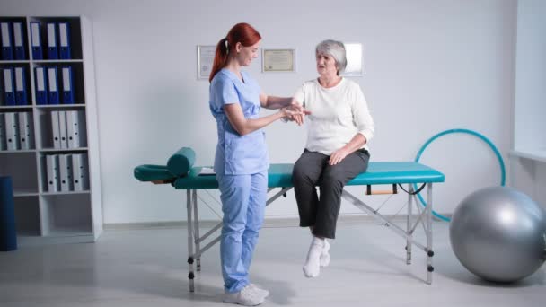 Врач в синей форме осматривает пациента на диване в офисе, пожилая женщина консультируется с физиотерапевтом с болью в руках - Кадры, видео