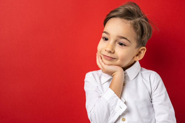 One boy male child five years old in front of red background wall wearing white shirt looking to the side leaning head on hand thinking contemplating copy space waist up - Photo, Image