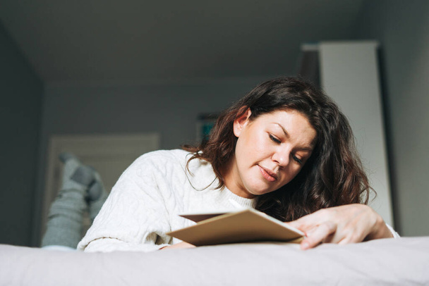Fiatal felnőtt barna nő negyven év plusz méret test pozitív fehér kötött pulóver írja jegyzetek tervező az ágyon otthon - Fotó, kép