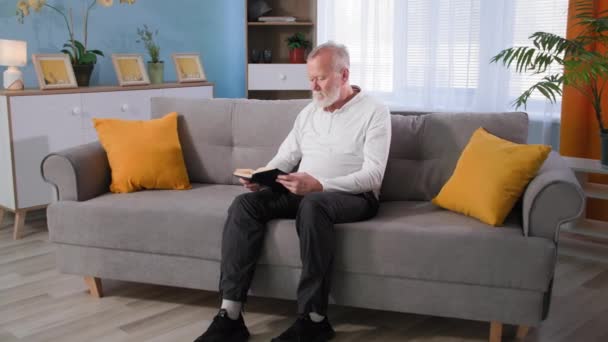 Vanha mies lukee kirjaa sitten nousee sohvalta ja tuntee kipua jalassaan, isoisä osteoporoosi hieroo polveaan kotona - Materiaali, video