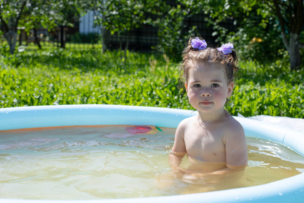Malá běloška 2 roky se koupe v nafukovacím bazénu na zahradě, dívka s mokrými vlasy sedí v bazénu a dívá se do kamery - Fotografie, Obrázek