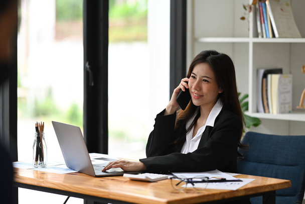 Mujer emprendedora sonriente trabajando con laptop y hablando por teléfono móvil. - Foto, Imagen