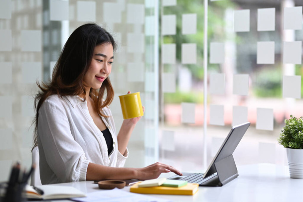 Очаровательная деловая женщина пьет кофе и проверяет электронную почту на планшете. - Фото, изображение