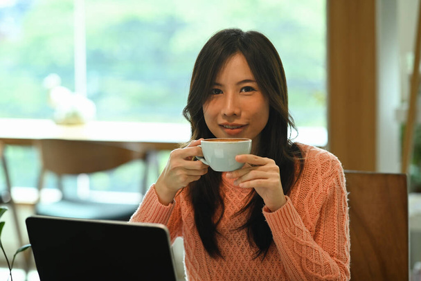 Nuori aasialainen nainen tilalla kahvikuppi ja katsot kameraa. - Valokuva, kuva