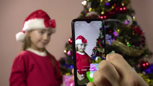 Noel Baba gibi giyinmiş küçük bir kız Noel ağacını süslüyor ve akıllı telefondan çekim yapmak için poz veriyor.. - Video, Çekim