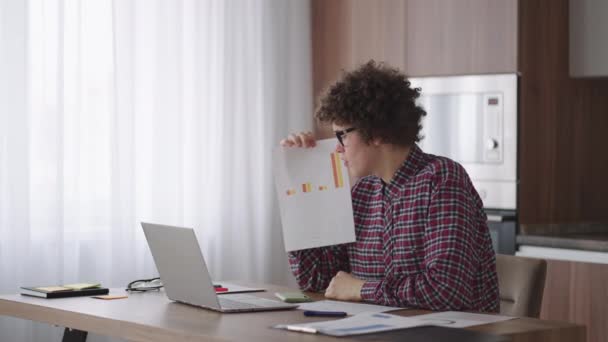 Curly - vlasy s brýlemi obchodník sedí v kanceláři z domova, dívá se do kamery a ukazuje na tablet s finančními informacemi zobrazeny v grafické podobě sloupcový graf - Záběry, video