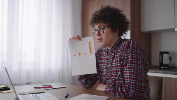 Curly - haired with glasses business man sitting at office from home desk looking at camera and pointing at a tablet with financial information displayed in graphical form column graph - Кадры, видео