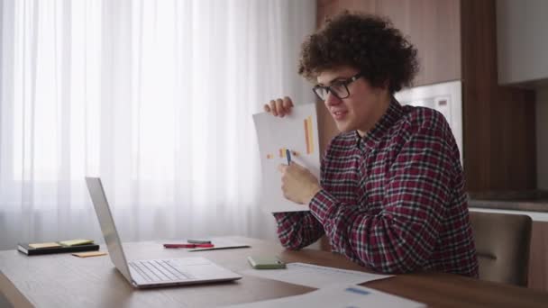 Curly - peludo con gafas hombre de negocios sentado en la oficina desde el escritorio de casa mirando a la cámara y apuntando a una tableta con información financiera que se muestra en el gráfico de forma gráfica columna - Metraje, vídeo