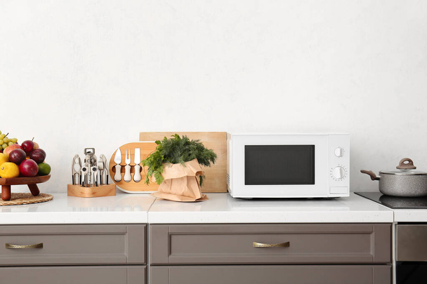Moderno horno microondas, utensilios de cocina y comida en el mostrador cerca de la pared de luz - Foto, Imagen