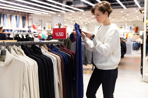 Молодая красивая брюнетка с короткой стрижкой в белом свитере выбирает стильную одежду в магазине в торговом центре - Фото, изображение