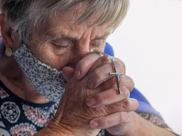 Virüse karşı koruyucu maskeli yaşlı bir kadın gözleri kapalı ve elleri bağlı dua ediyor. Hıristiyanlık kavramı - Fotoğraf, Görsel