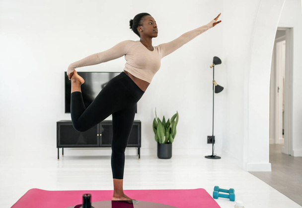 Młoda czarnoafrykańska kobieta stojąca i rozciągająca się na macie do jogi, w nowoczesnym salonie w domu. Postawa jednej nogi - Zdjęcie, obraz
