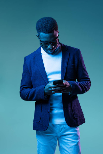 Half-length portrait of young stylish african man, student wearing white neck-polo and jacket isolated on blue background in neon light. - Foto, Imagem