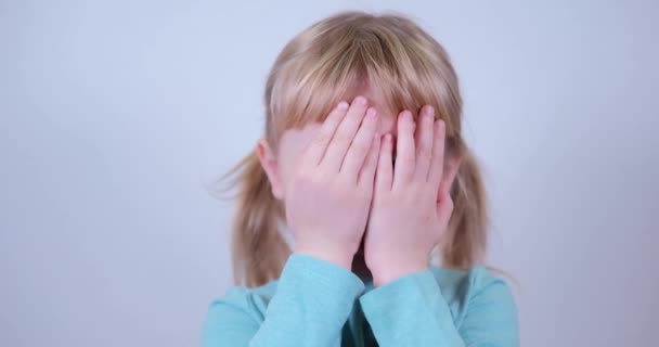 Söpö pieni vaalea tyttö peittää kasvonsa käsillään sisätiloissa. Leikkii piilosta, kurkistaa buuaa. - Materiaali, video