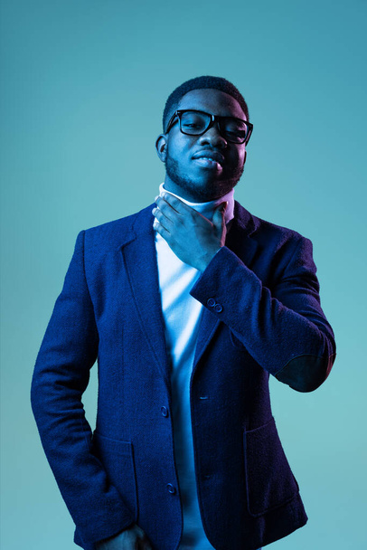 Half-length portrait of young stylish african man, student wearing white neck-polo and jacket isolated on blue background in neon light. - Photo, Image