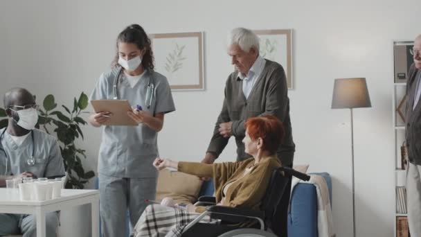 Mittlere Aufnahme zweier multiethnischer medizinischer Mitarbeiter, die die Temperaturen von Senioren im Pflegeheim messen und ihnen Tabletten in Einwegbechern geben - Filmmaterial, Video