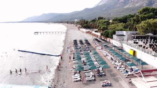 Vista aérea de una playa blanca con filas de tumbonas con vacacionistas y una costa de montaña con hoteles en una pequeña ciudad - Metraje, vídeo