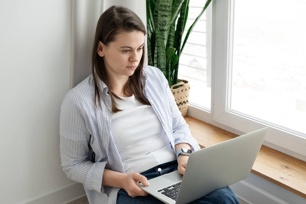 Middle-aged woman working from home. Girl using laptop at home, sitting near a large window. Remote work concept - Foto, Imagem