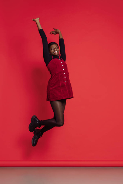 Full-length portrait of happy African girl in casual clothes jumping isolated on red studio background. Concept of emotions, holidays - Φωτογραφία, εικόνα