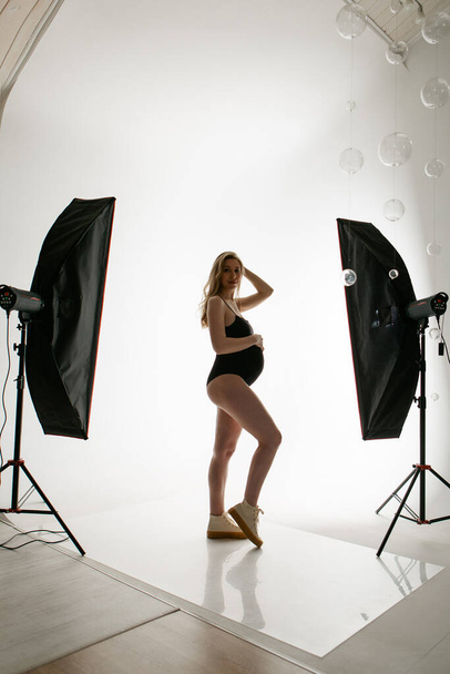 a pregnant woman in a black suit strokes her belly on a white background. A girl in the studio in the spotlight. professional lighting. - Foto, Imagem