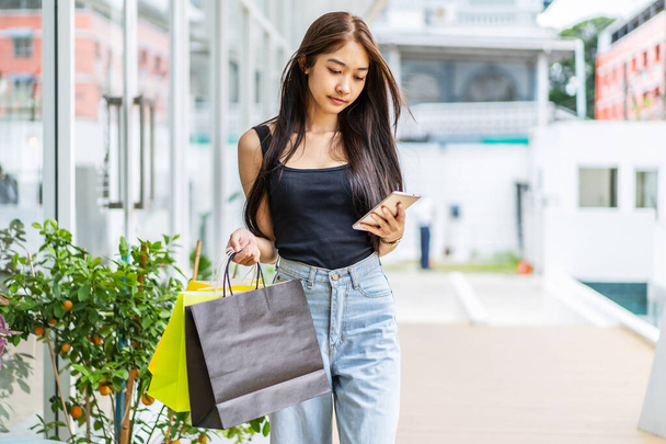 Asian girl is holding shopping bags, using a smartphone while doing shopping in the mall, shopping concept, portrait - Фото, зображення