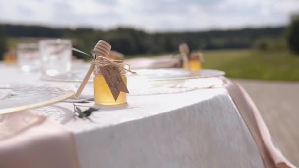 Ухоженный открытый стол со стеклянной посудой и маленькими банками для меда с деревянными медовыми ковшками для гостей свадьбы. Размытый фон природы - Кадры, видео