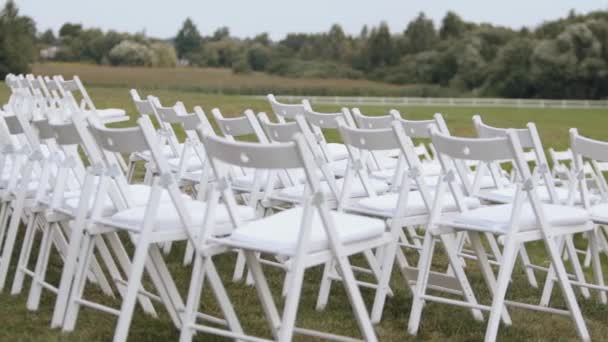 Rows of white chairs on a green lawn surrounded by trees for guests at a wedding ceremony - Filmati, video