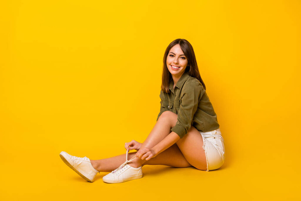 Photo of young lady sit floor tie shoelace wear green shirt krótkie trampki izolowane żółty kolor tło. - Zdjęcie, obraz