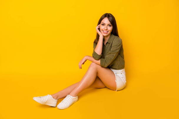 Photo of cheerful young lady sit floor hand face wear green shirt mini skirt footwear isolated yellow color background - Фото, изображение
