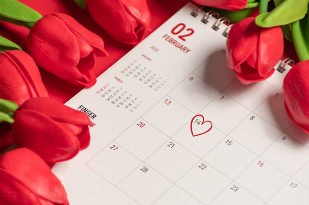 El corazón de la marca en el 14 calendario de febrero con tulipanes rojos de flores en el fondo rojo en un escritorio, el concepto para el día de San Valentín el 14 de febrero de 2022. Primer plano, enfoque selectivo, fondo borroso - Foto, Imagen