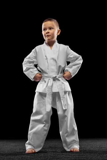 One little kid, boy, taekwondo athlete wearing white dobok isolated over dark background. Concept of sport, education, skills - Photo, Image
