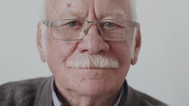 Langzaam close-up portret van blanke senior man met grijze snor in bril glimlachend naar camera op witte achtergrond - Video