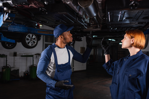 Side view of interracial mechanics pointing at auto in car service  - Foto, Imagem